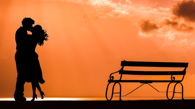 a couple kissing standing next to a bench on a beach at sunset