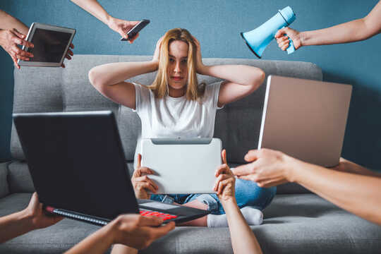 Woman sits with hands over her ears as megaphone, cell phone, 2 laptops, 2 iPads are thrust in her face